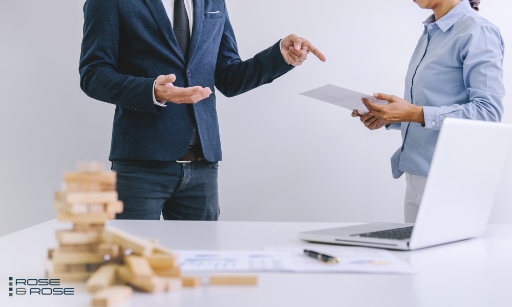 Two business professionals, one pointing at a document held by the other, standing by a desk with a laptop and a tumbling tower game.